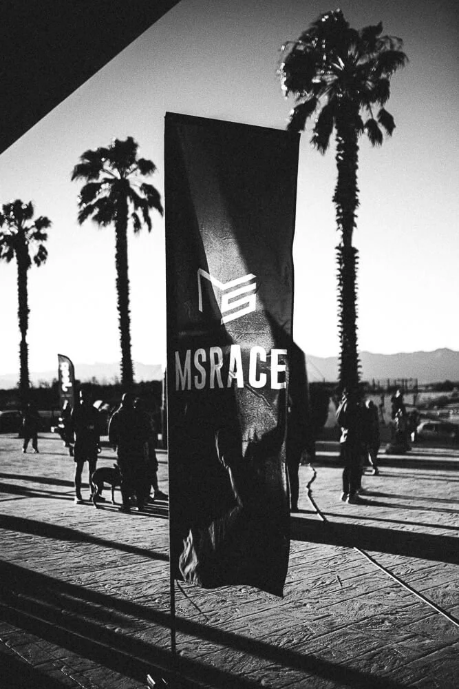 Black and white outdoor scene featuring a vertical banner with the text 'MSRACE' and a stylized logo, palm trees, and several people standing near parked cars in the background.