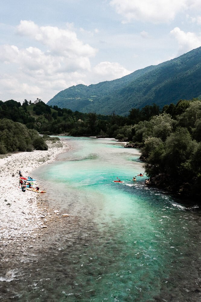 A scenic river flowing through a lush green valley with mountains in the background, some people on kayaks and paddleboards on the river, and a few others relaxing on the rocky shore.