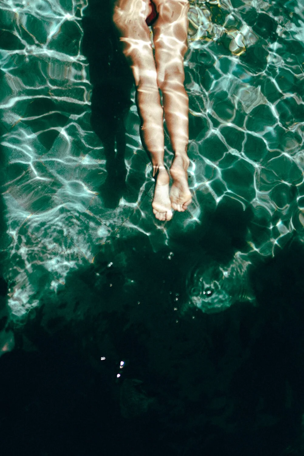 A person floating in a pool with their legs extended, sunlight creating ripples and reflections on the water.