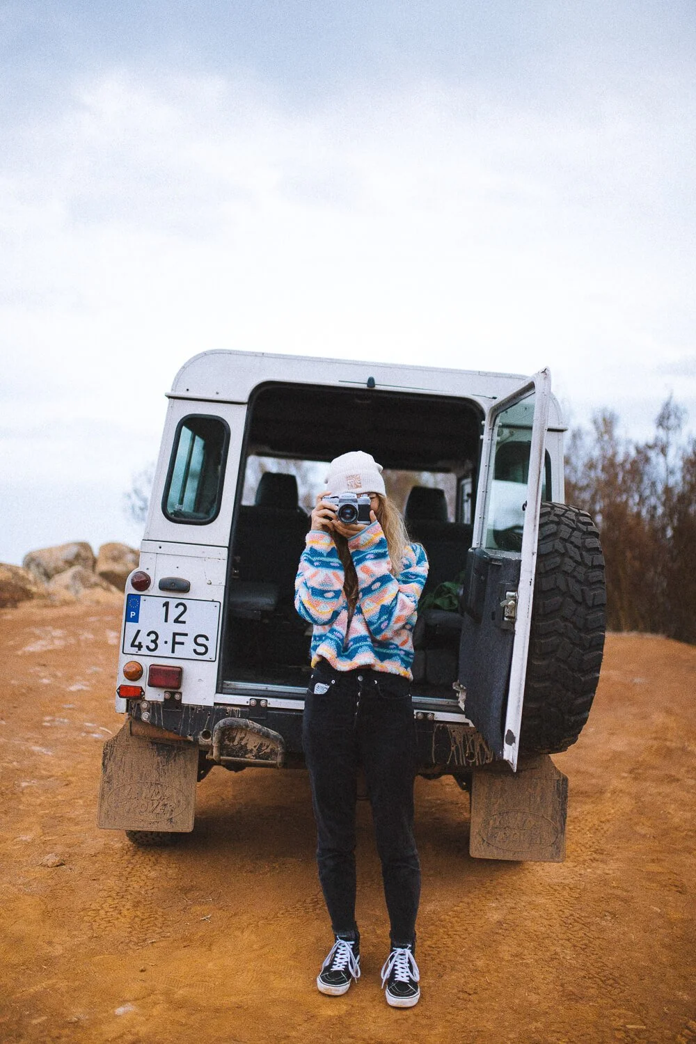 A woman standing outdoors on dirt ground, taking a photo with a camera. She is wearing a white beanie, colorful sweater, black pants, and sneakers. Behind her is an open-back white vehicle with a spare tire on the back, license plate reading '12 43-F