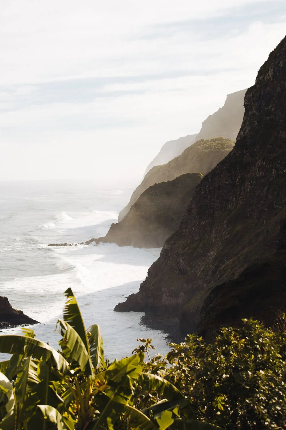 Coastal cliffs with lush greenery in the foreground and ocean waves crashing below, under a cloudy sky.