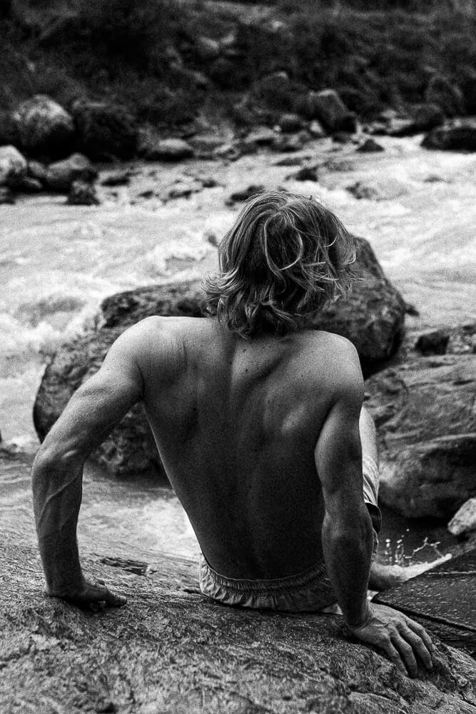 A shirtless man with long hair sitting on a rock near a river.