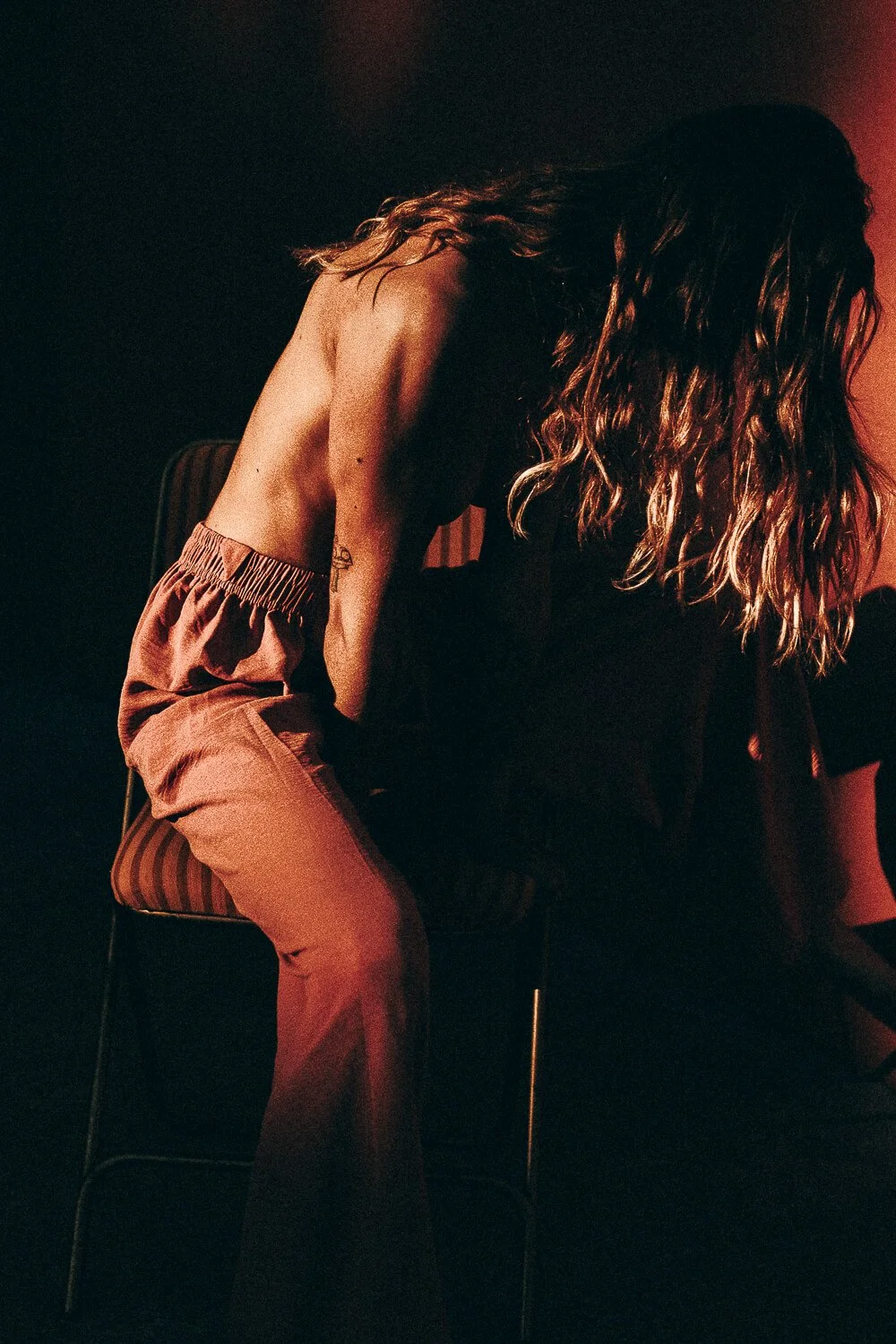A woman with long wavy hair is sitting on a chair, facing downward, in a dimly lit room with red lighting.