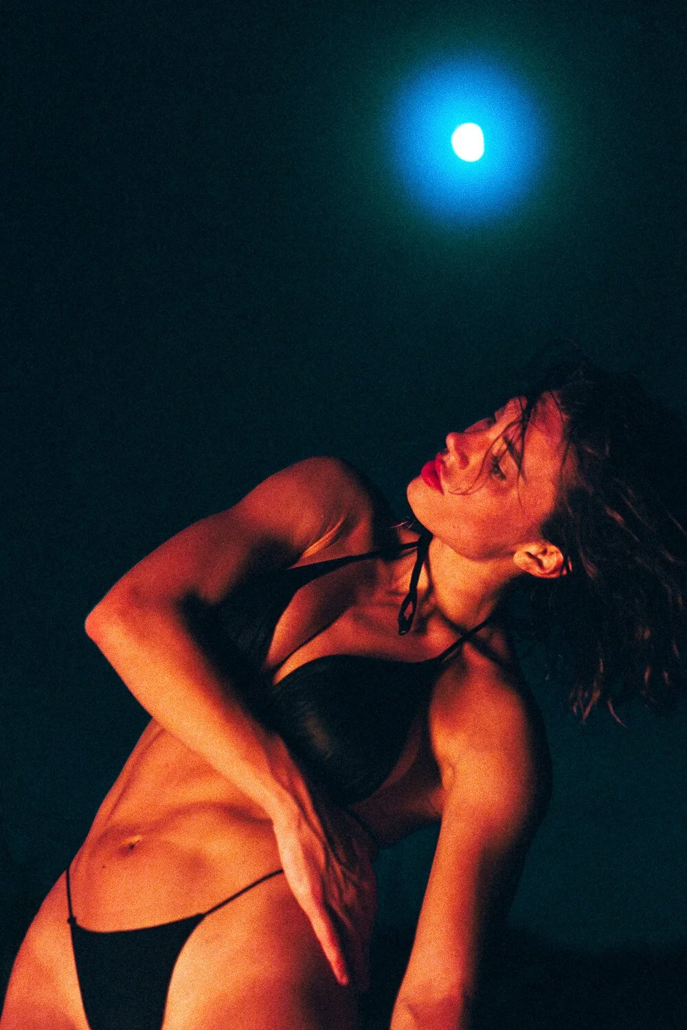 A woman in black lingerie and red satin shorts lying down in a dark room with a blue moon in the background.