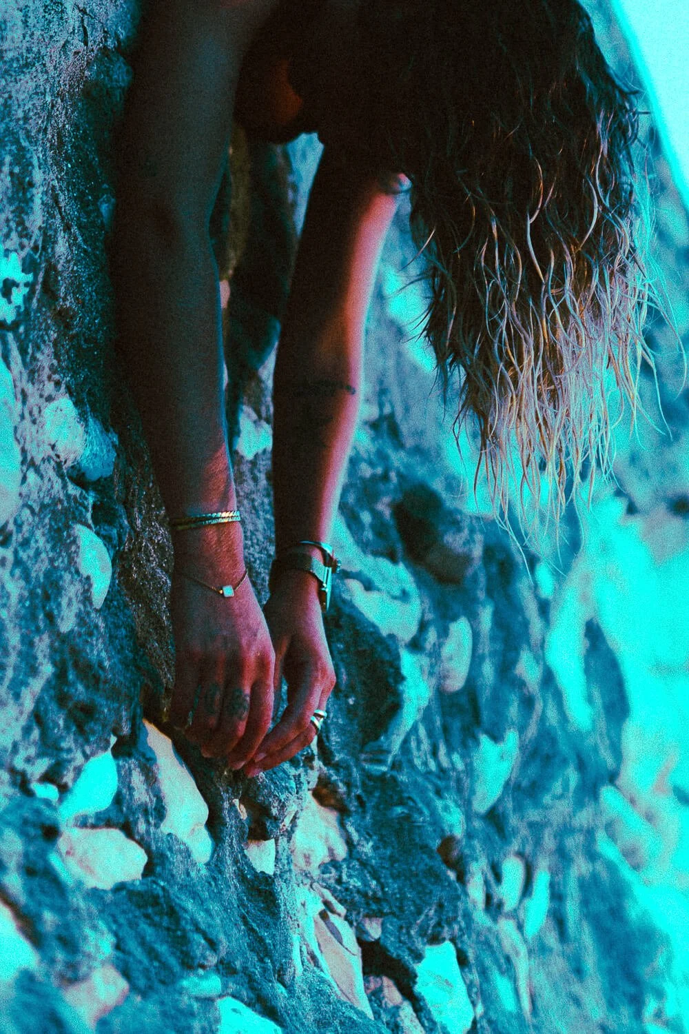 A person with long, curly hair and jewelry on their arms and fingers is leaning over a rocky surface, holding onto the rocks with both hands, in a setting with blue lighting.