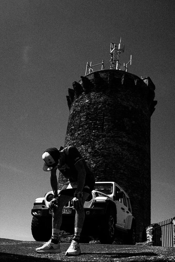 A person wearing a helmet and athletic gear is standing and bending down in front of a white SUV, with a tall stone tower with antennas on top in the background.