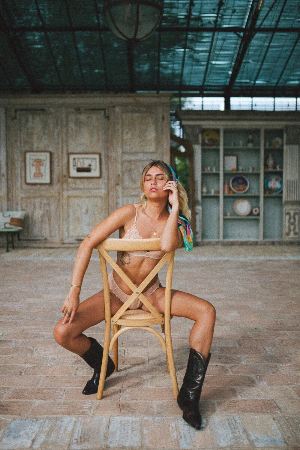 A woman wearing lingerie, black boots, and a colorful headscarf, seated on a wooden chair with legs apart in an indoor space with a rustic, vintage decor background.