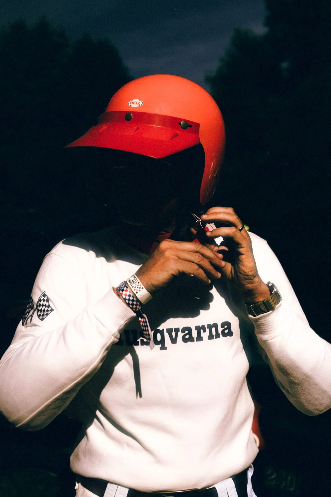 A person wearing a red motorcycle helmet with a Bell sticker, adjusting or putting on a black face mask. The person is wearing a white long-sleeve shirt with black text and checkered pattern details on the sleeves and wristbands.