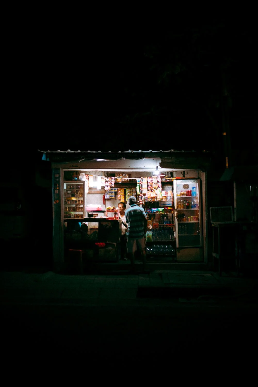 Une petite boutique éclairée la nuit avec deux personnes à l'intérieur, vendant divers produits comme des snacks, boissons et autres articles de commerce.