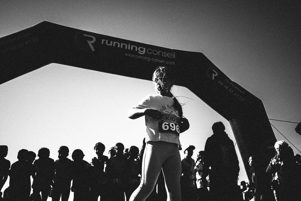 A young female runner adjusting her watch under a large archway at a race event, with other runners and spectators in the background, in black and white.