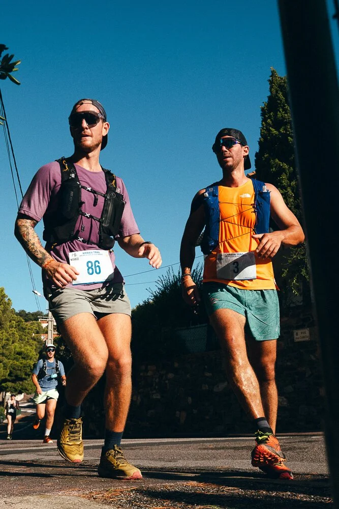 Two male runners participating in a race outdoors, wearing athletic gear and sunglasses, with bib numbers 86 and 3.