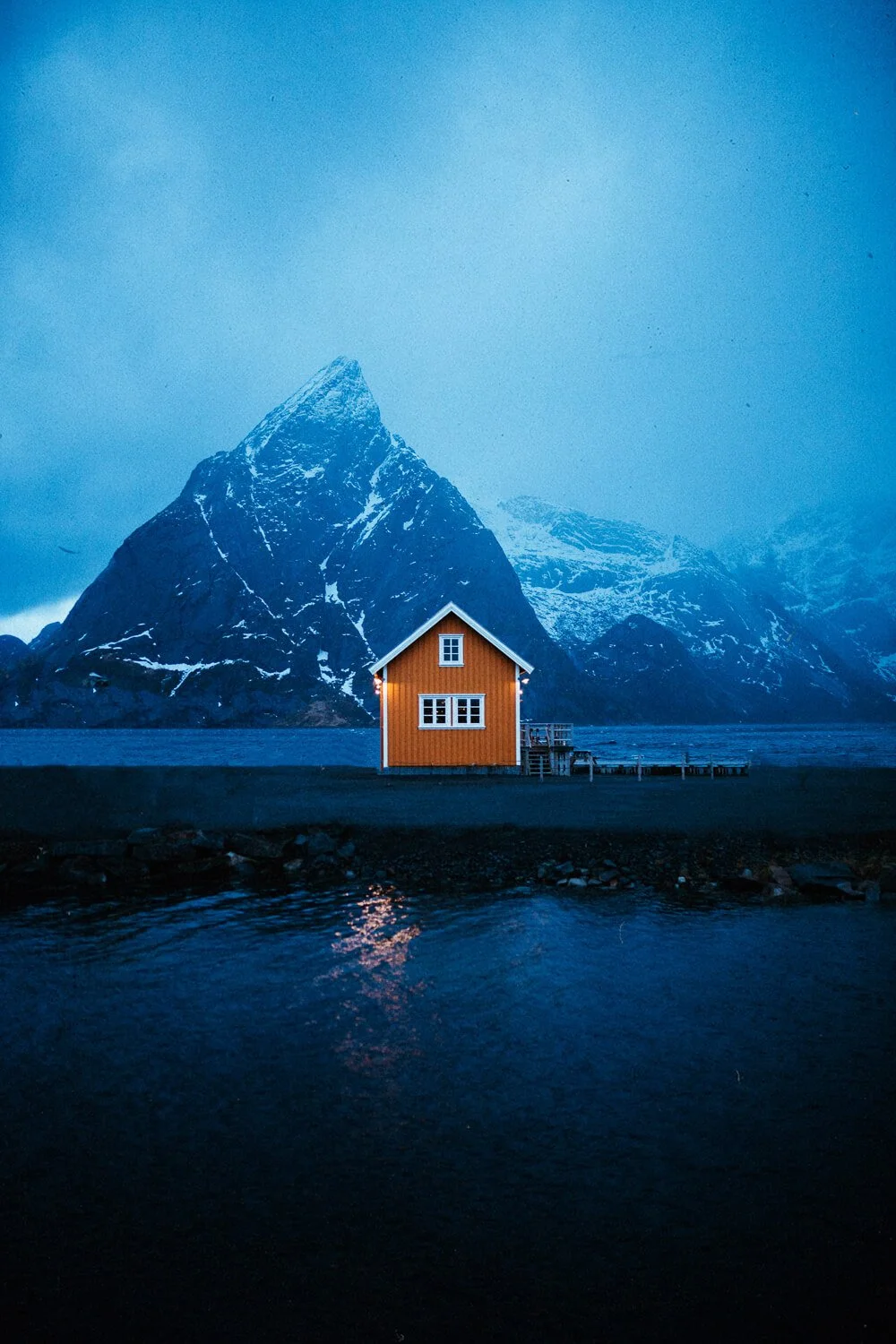 Une cabane en bois orange située au bord de l'eau, avec des montagnes enneigées en arrière-plan, sous un ciel bleuâtre.