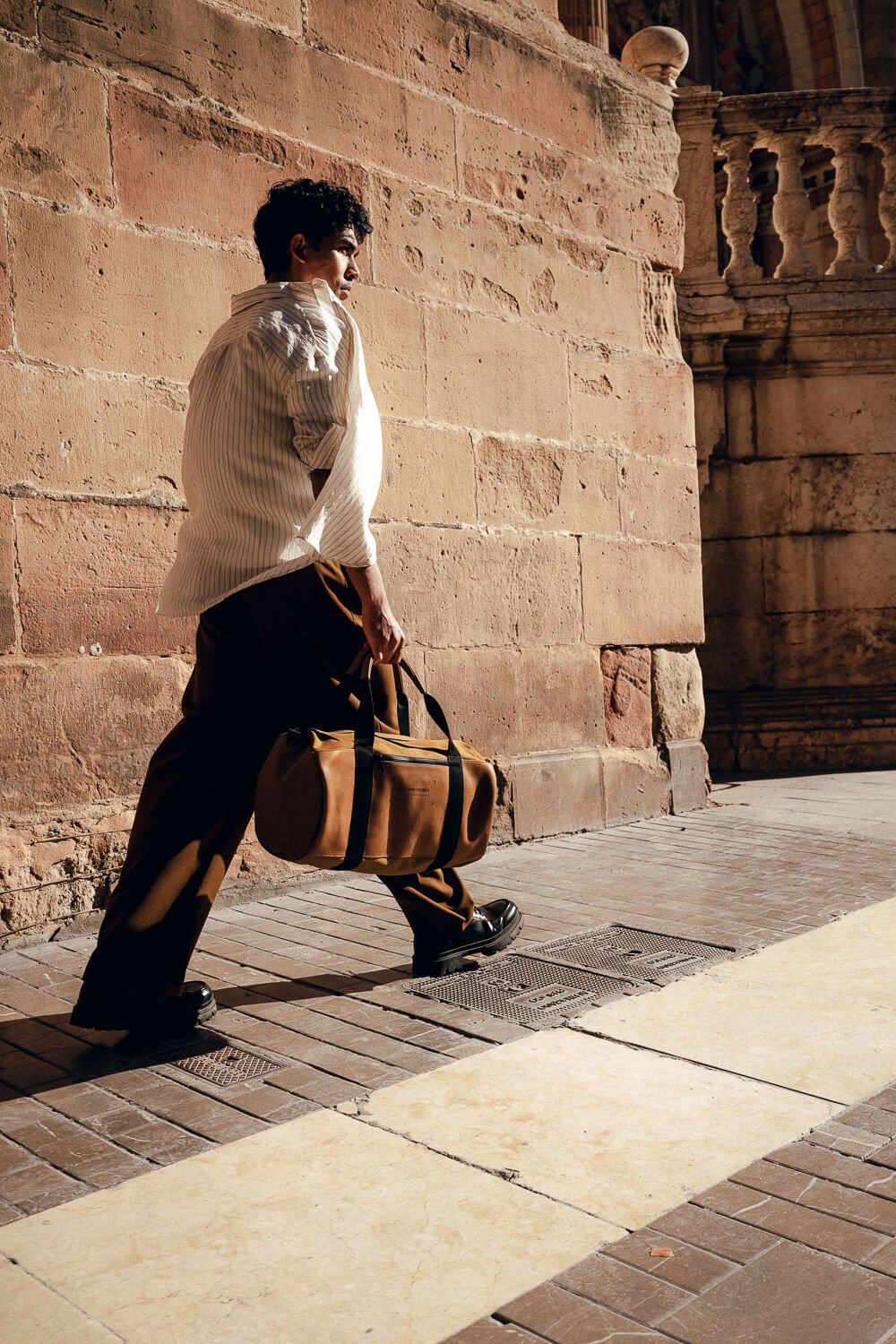 A man with dark, curly hair wearing a light-colored shirt, dark pants, and black shoes, walking on a sidewalk next to a brick wall, carrying a brown duffel bag.