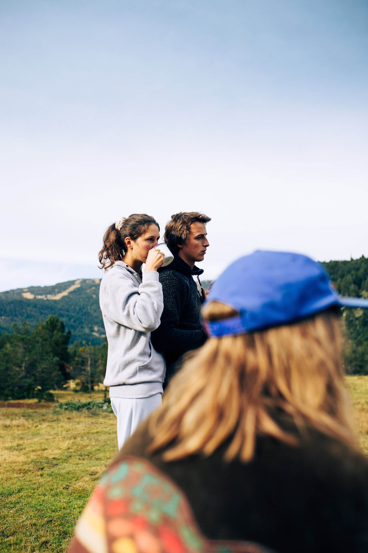 Trois personnes en plein air, dont une femme buvant dans une tasse, avec un paysage montagneux en arrière-plan.