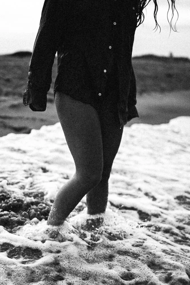 Person standing in shallow ocean water, wearing a dark wet shirt and shorts, with waves around their legs, on a beach with rocky terrain in the background, in black and white.