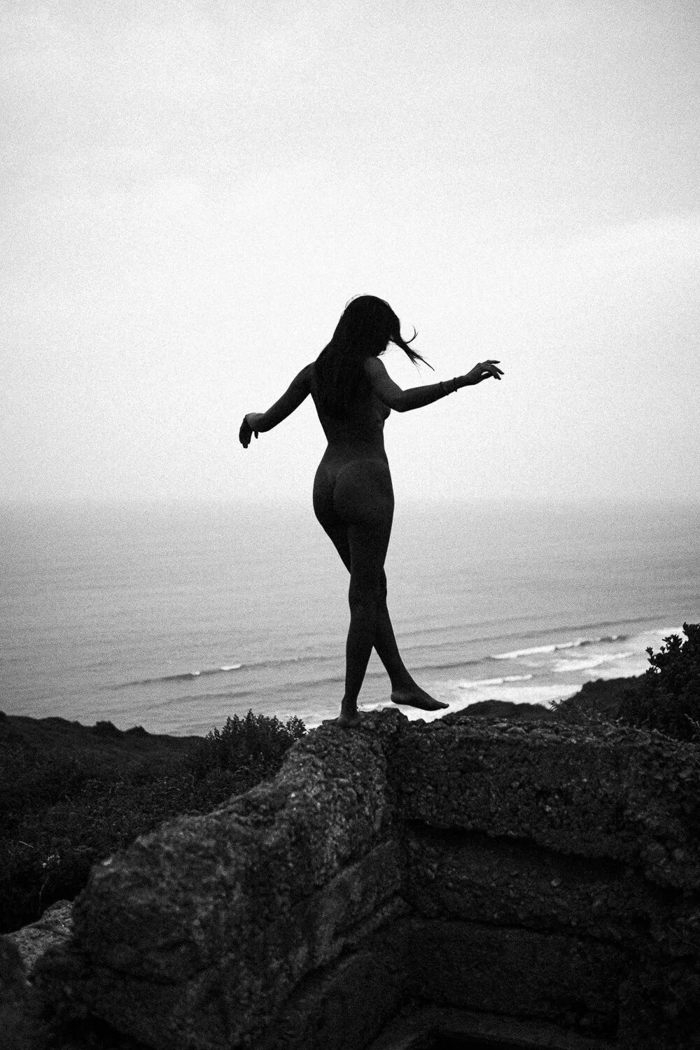 Silhouette of a woman standing on a rock overlooking the ocean, with arms outstretched and hair flowing in the wind, in black and white.