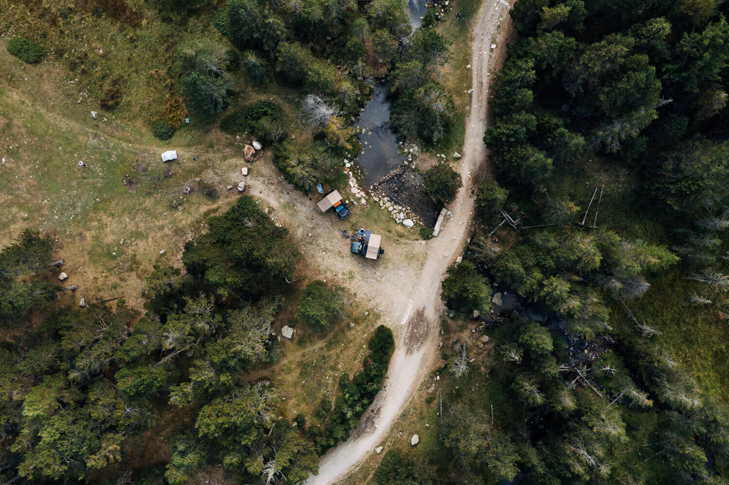 Vue aérienne d'un camping dans une forêt avec une petite route, une rivière, des véhicules et des arbres.