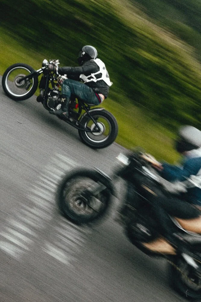 Two motorcyclists riding on a road, with one in focus and the other blurred, indicating motion.