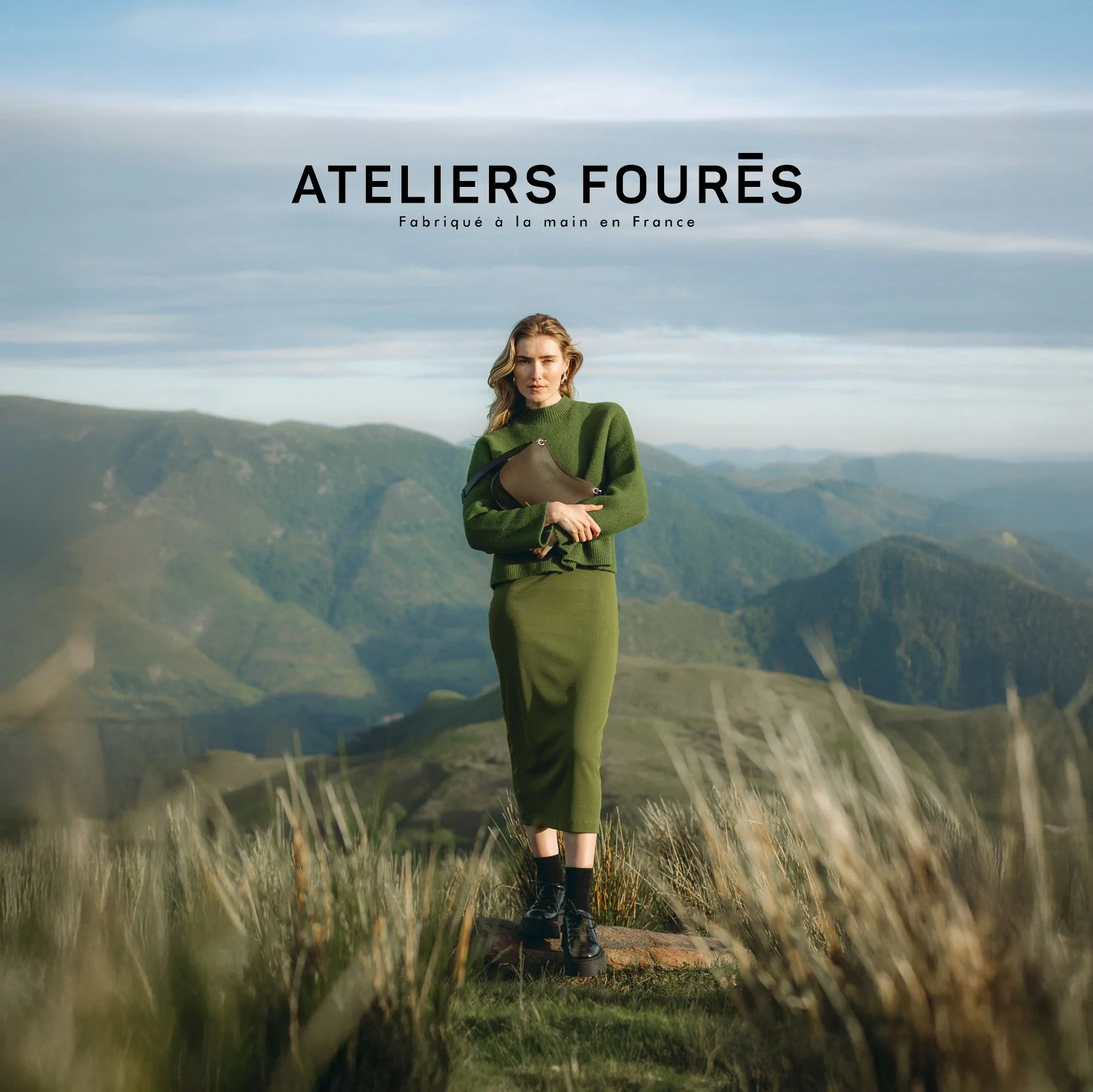 Une femme en vêtements verts debout dans un paysage montagneux avec un ciel nuageux, tenant un sac à main, pour une publicité de vêtements faits à la main en France.