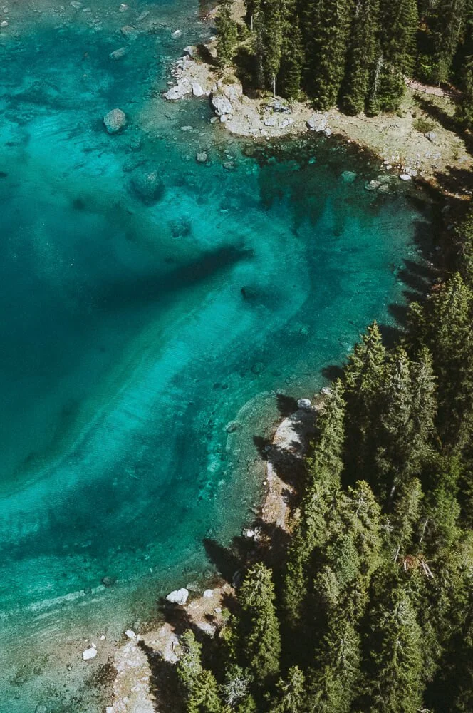 Aerial view of a clear, turquoise lake surrounded by dense green trees and rocky shoreline near a forest.