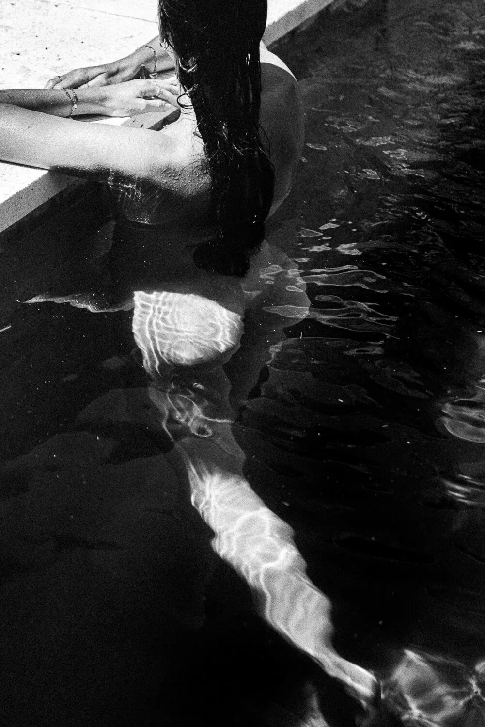 A person with long hair leaning against the edge of a pool, with their back to the camera, partially submerged in the water.