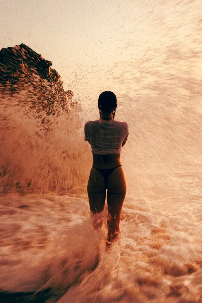 Silhouette of a woman with her back to the camera, standing in the ocean with a large wave crashing behind her during sunset.