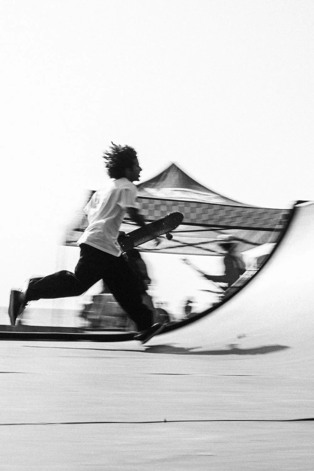 A person running on a skateboard in black and white, holding a skateboard under their arm, with a tent and other people in the background.
