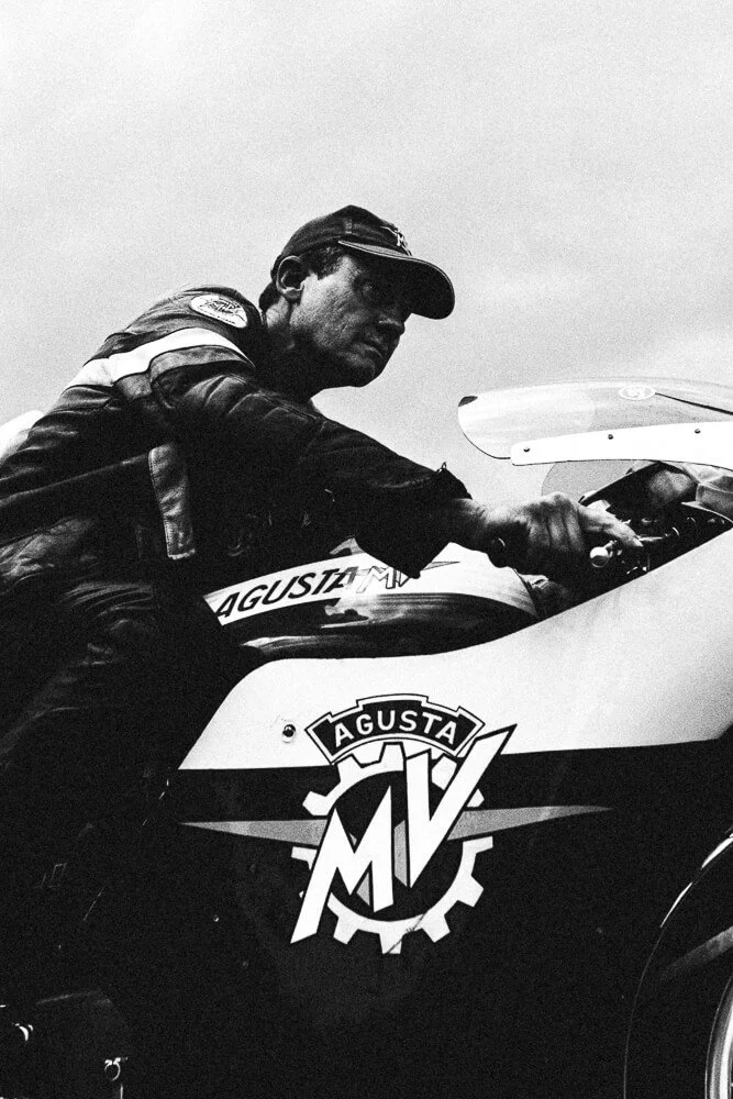 A man in a racing suit and cap leaning on a motorcycle, with the "Agusta MV" logo visible on the bike.