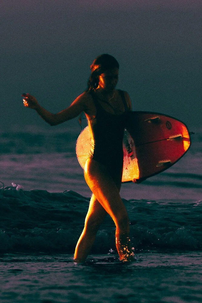 A woman holding a surfboard walking in the ocean at sunset.