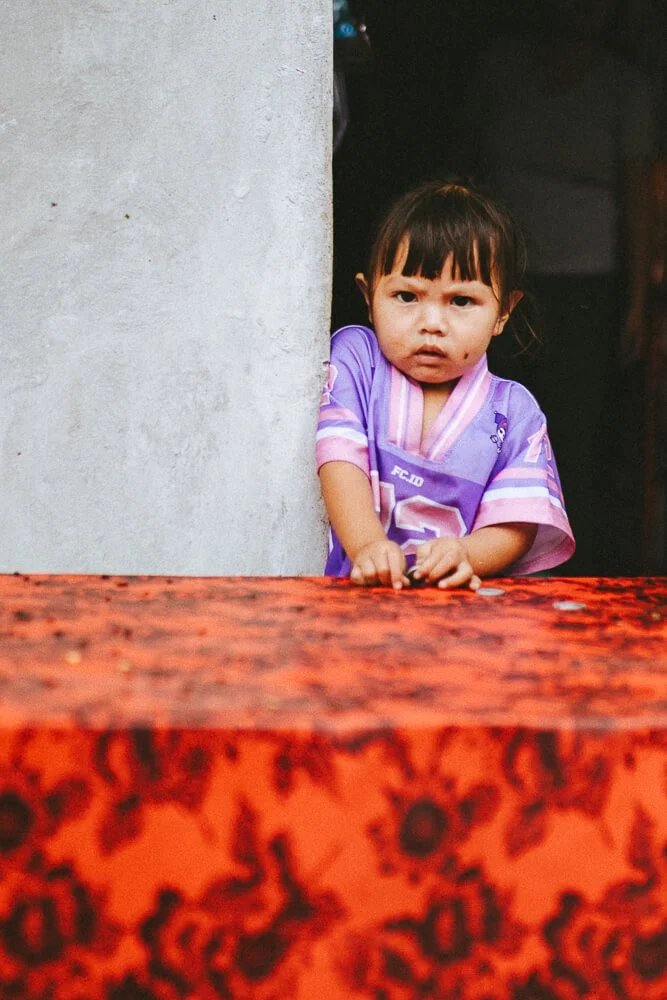 A young girl with dark hair and bangs, wearing a purple sports jersey, peeks out from behind a wall with a curious or concerned expression.