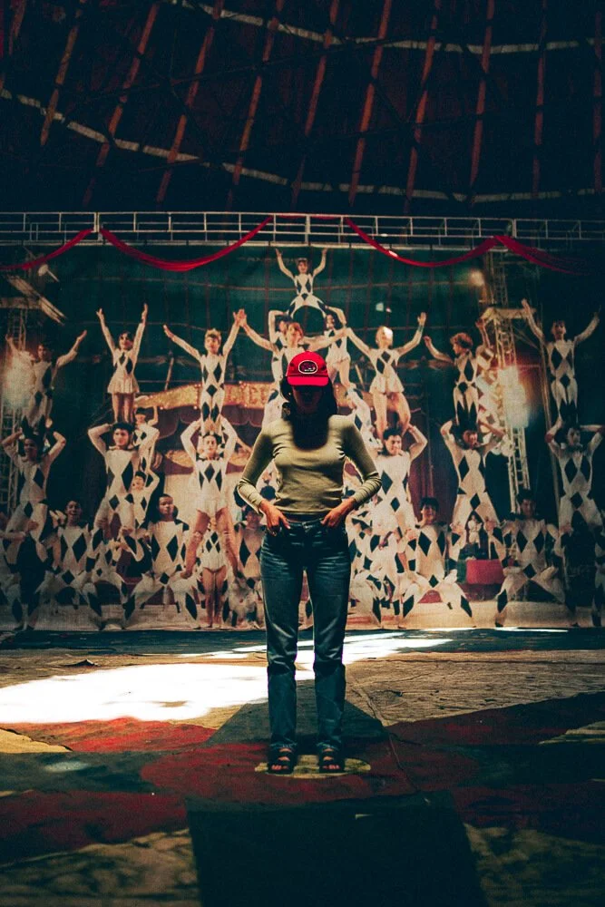 Person standing with head bowed and hands in pockets in front of a circus or performance backdrop with performers in acrobatic poses.