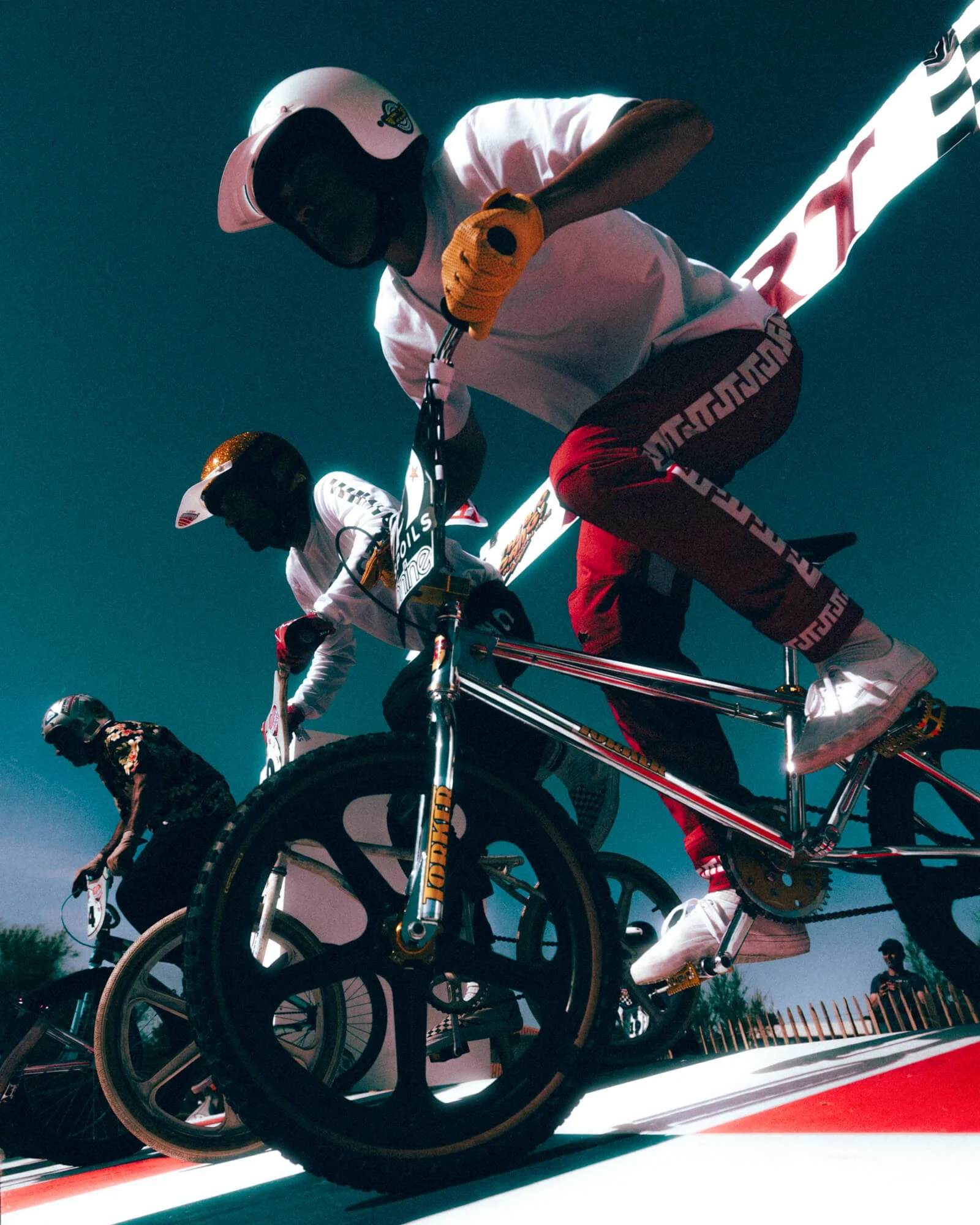 Cyclists riding on a race track during a race, with one cyclist in foreground wearing a white helmet and white jersey, and others behind him.