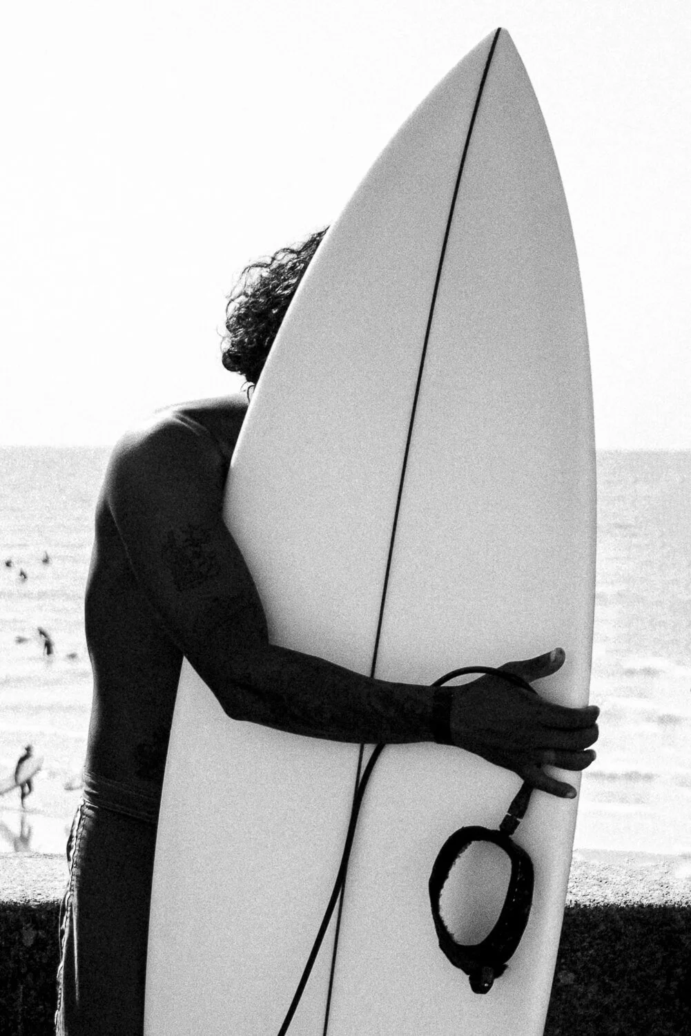 A person holding a surfboard at the beach with several people in the background near the water, black and white photo.