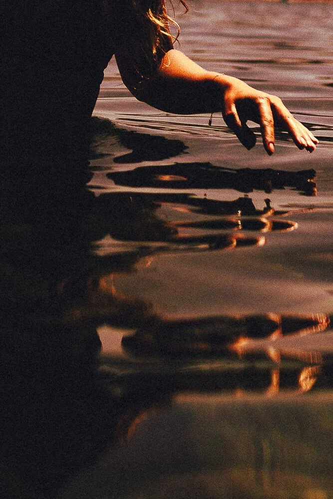 A person with long hair reaching into a body of water during sunset, with their reflection visible on the surface.