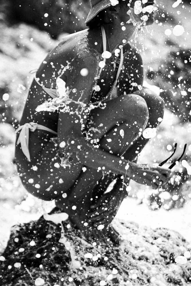 Person crouching in water with splashes, wearing a hat, sunglasses, and a swimsuit, holding a rake or similar tool.