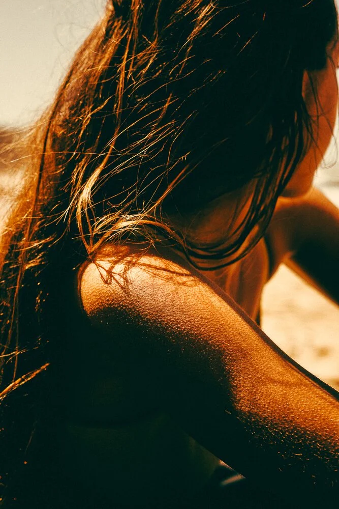 Close-up of a woman's shoulder and hair illuminated by warm sunlight, with a sandy beach in the background.