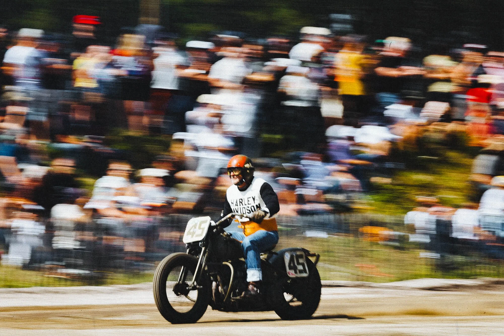 Un motard portant un casque orange et un gilet avec l'inscription Harley Davidson, en train de faire une course sur une piste, avec une foule de spectateurs flous en arrière-plan.