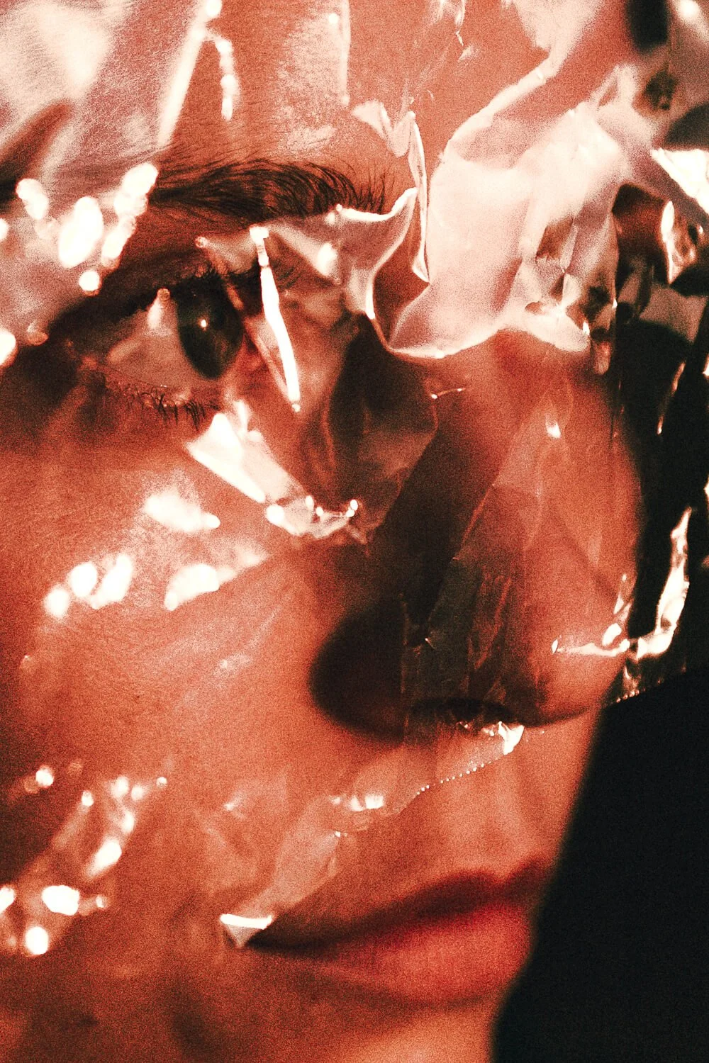 Close-up of a woman's face with dark eye makeup, partially covered by transparent plastic with white petals reflecting light.