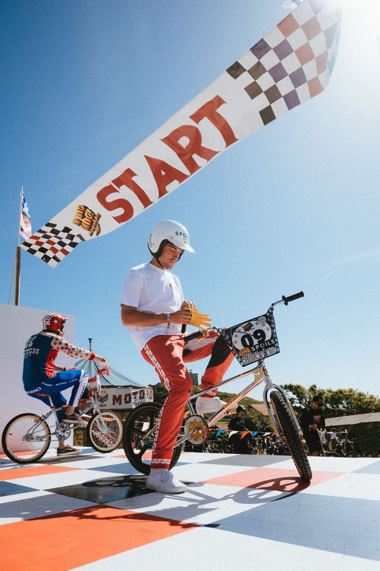 A person in a helmet and sportswear performing a bike stunt during a cycling event, with spectators and other cyclists in the background, on a sunny day.