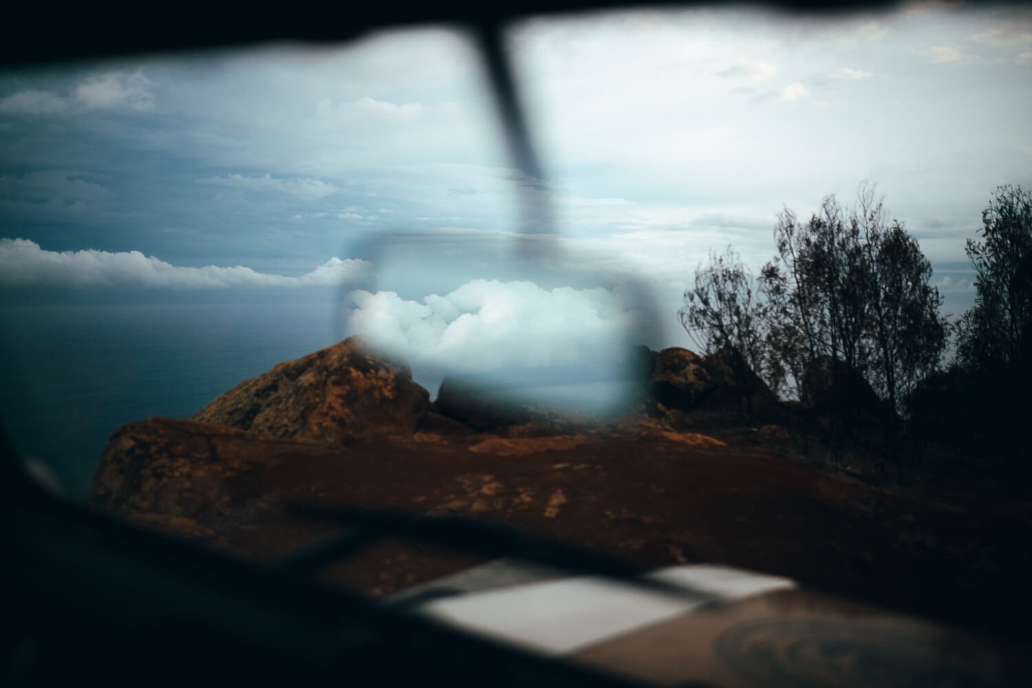 Mountain landscape with rocks, trees, and cloudy sky viewed through a vehicle's side mirror.