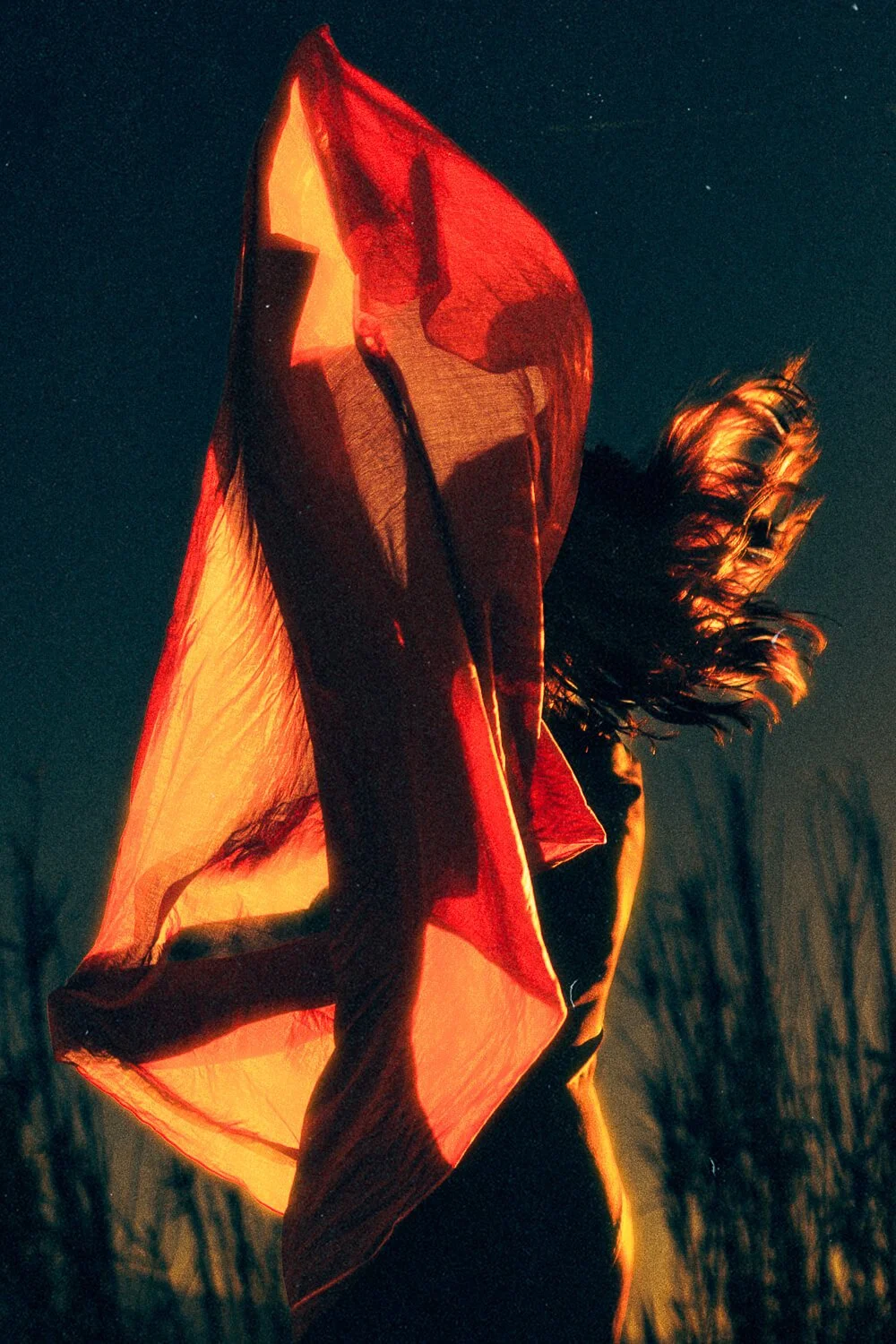 A woman outdoors at night with her hair blowing in the wind, wearing a red-orange cape illuminated from beneath, creating a glowing effect.