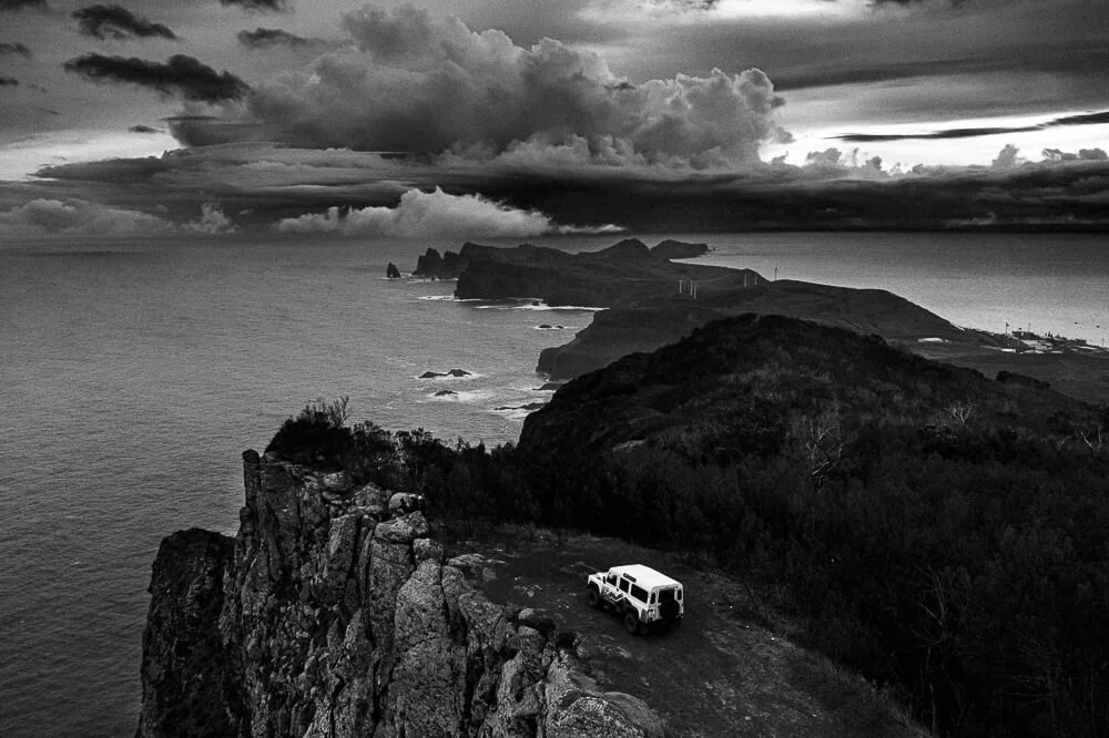 A black and white photo of a coastal landscape with cliffs, ocean, and a cloudy sky. A white vehicle is parked near the edge of the cliff.