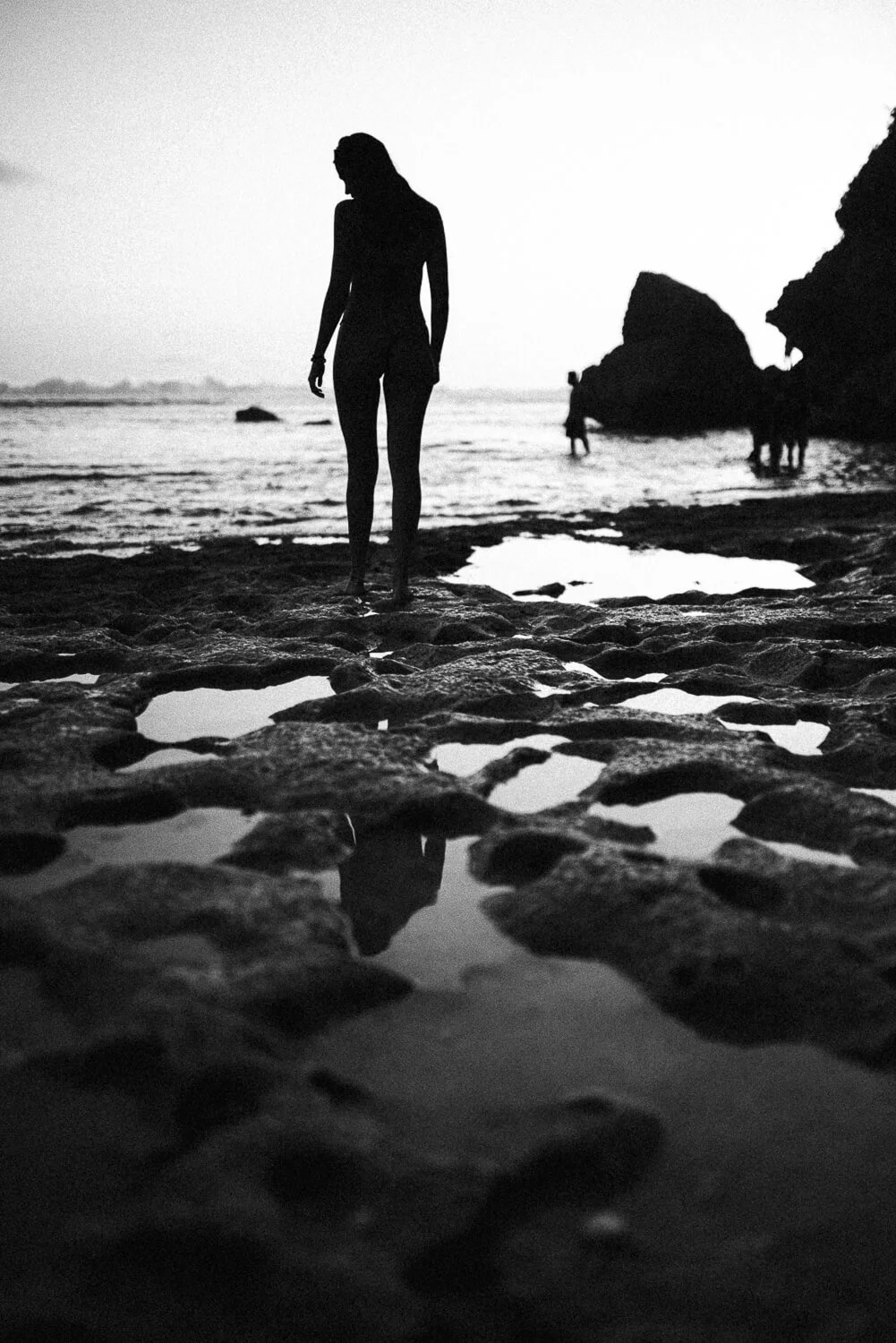 Silhouette of a woman walking on a rocky beach with ocean and distant people in the background, black and white photo.