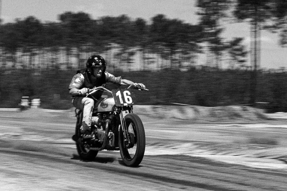Black and white photo of a motorcycle race with a rider wearing a helmet with stars, riding a vintage motorcycle numbered 16 on a dirt track, with trees blurred in the background.