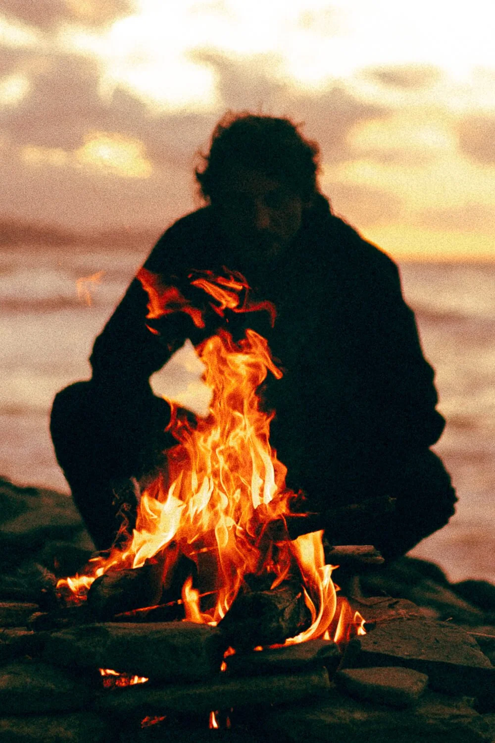 Person sitting by a campfire on a beach during sunset, with clouds in the sky.