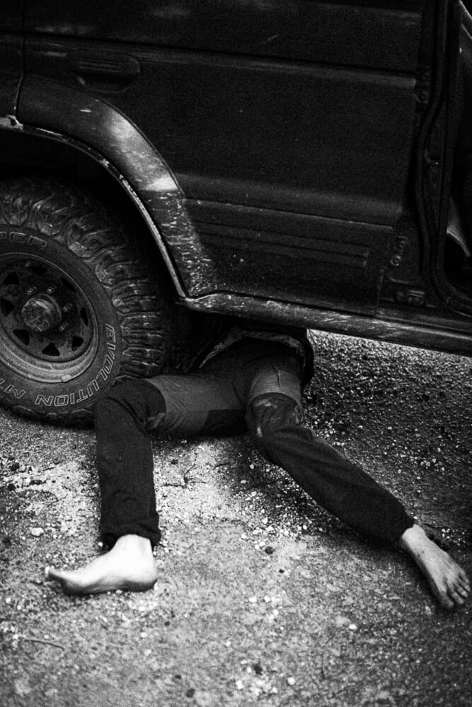 A person lying on the ground underneath a dark SUV, with only their legs and feet visible, on a gravel surface.
