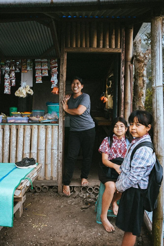 A smiling woman standing at the entrance of a small, rustic shop, with two young girls outside. The shop is made of bamboo and wood, with snacks and supplies visible inside. One girl is sitting on a small stool, and the other stands beside her, both 