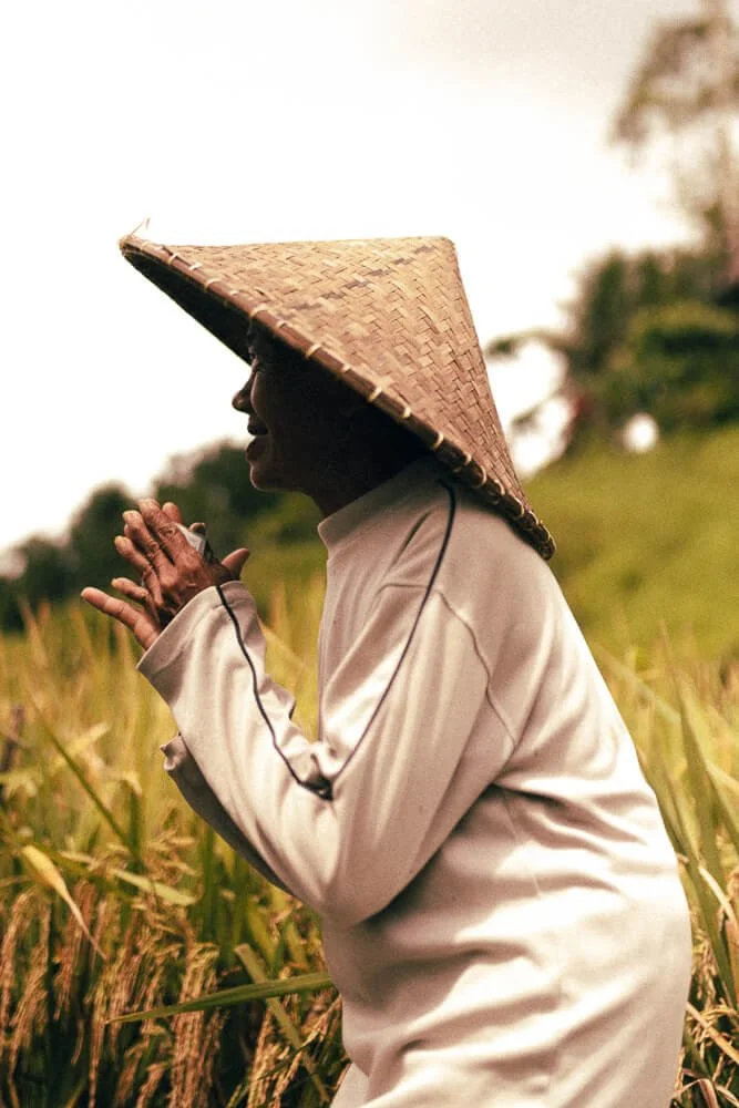 A person wearing a traditional conical straw hat and a white long-sleeve shirt standing in a field of tall grass or crops, with hands clasped together in a prayer-like position.