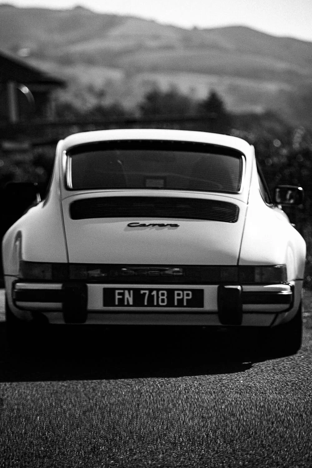 Black and white photo of the rear view of a classic Porsche Carrera car with a license plate reading 'FN 718 PP', parked outdoors with hilly landscape in the background.