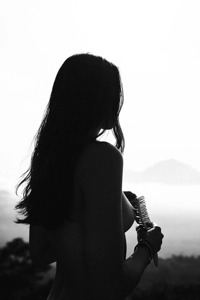 Silhouette of a woman with long hair holding a hairbrush and looking towards the horizon over water and distant mountains.
